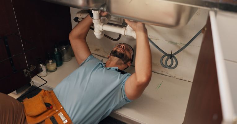An Atlas Trillo plumber wearing safety glasses is lying on the floor beneath a kitchen sink, working on the pipes.
